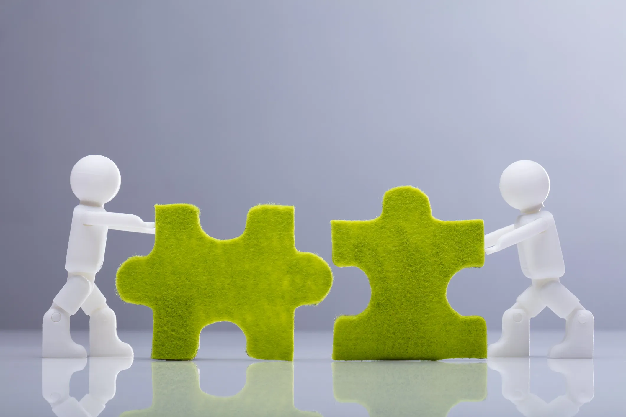 Side View Of Two Miniature Human Figures Solving Green Jigsaw Puzzles On Grey Background