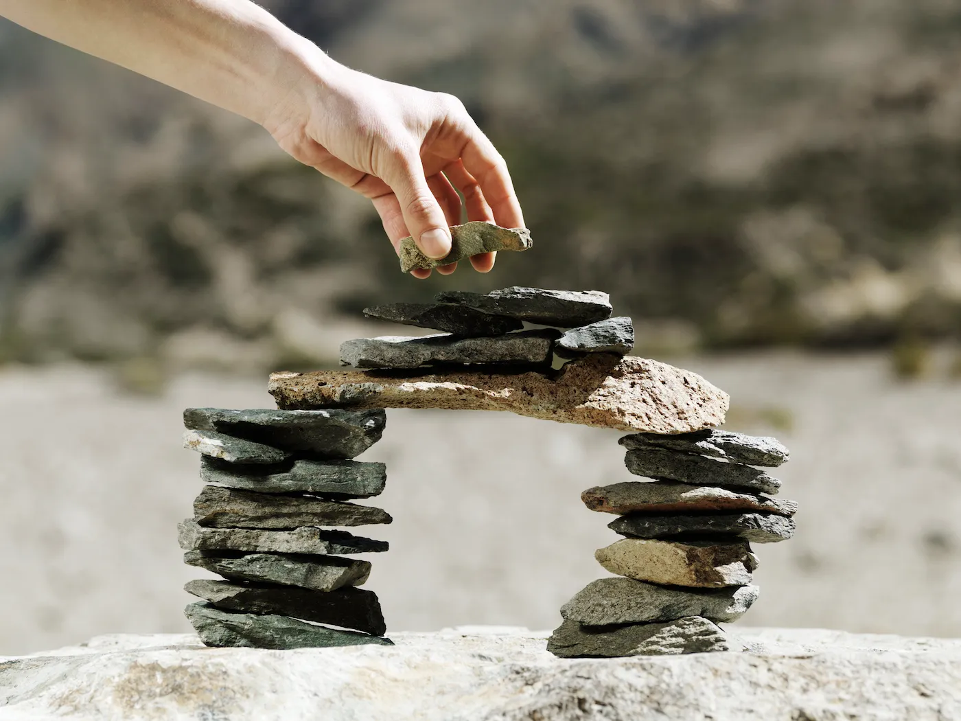 Final stone being placed by hand on a balancing miniature model bridge made of small flat rocks outside