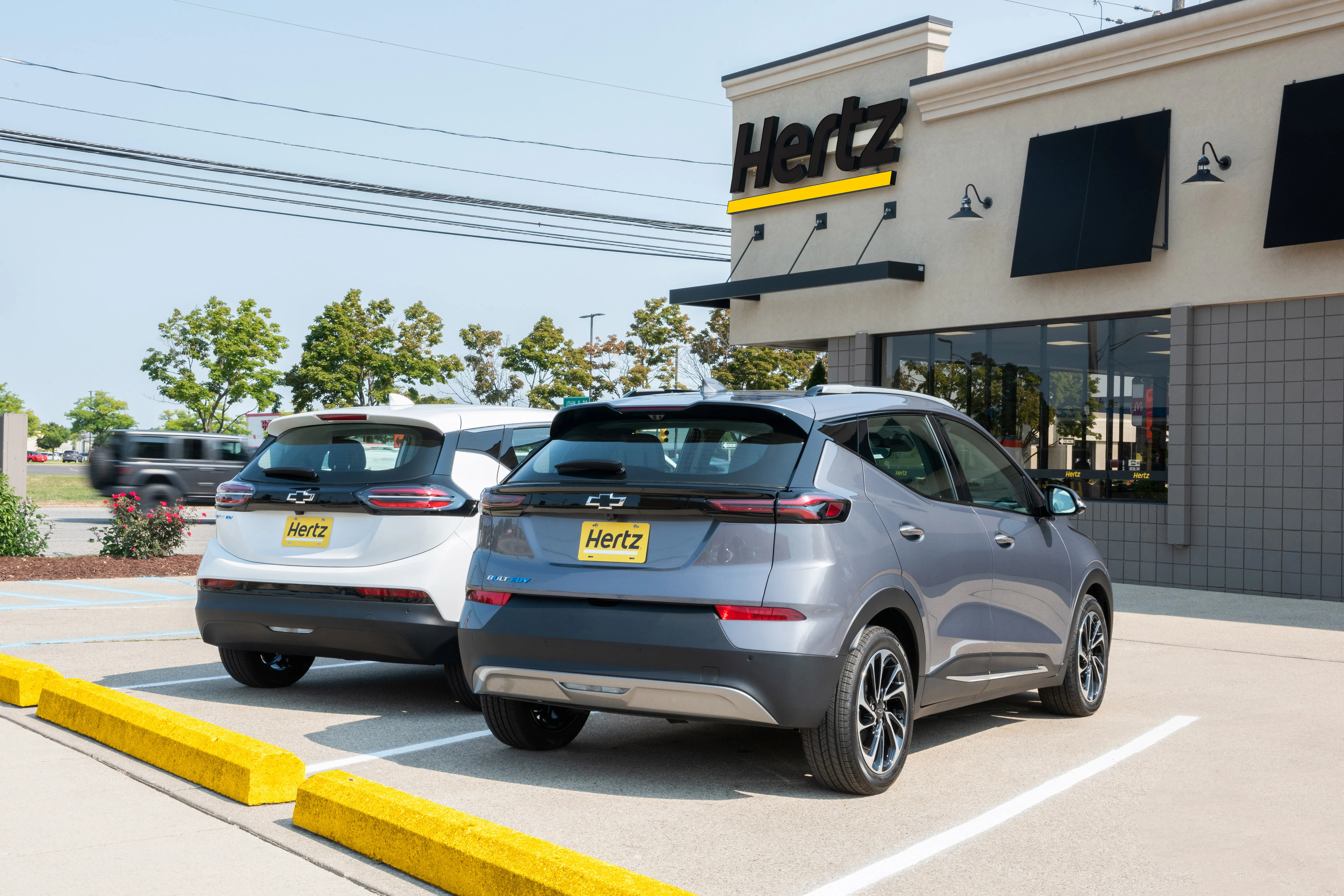 A Summit White 2023 Chevrolet Bolt EV and a Gray Ghost Metallic 2023 Chevrolet Bolt EUV parked in front of a Hertz rental location in Michigan. Hertz will begin taking deliveries of the Chevrolet Bolt EV and Bolt EUV in the first quarter of 2023.