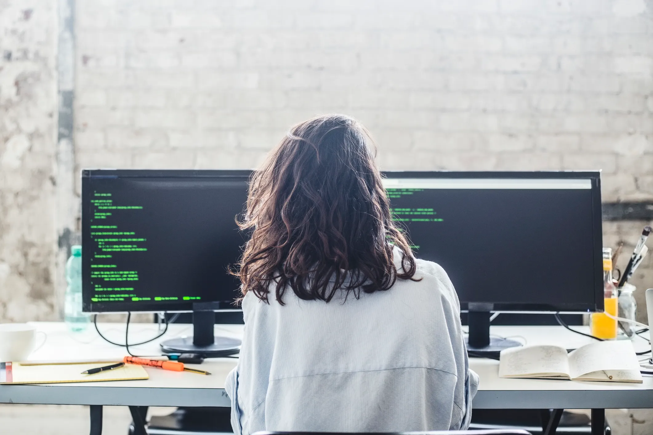 back of woman sitting at desk with two monitors