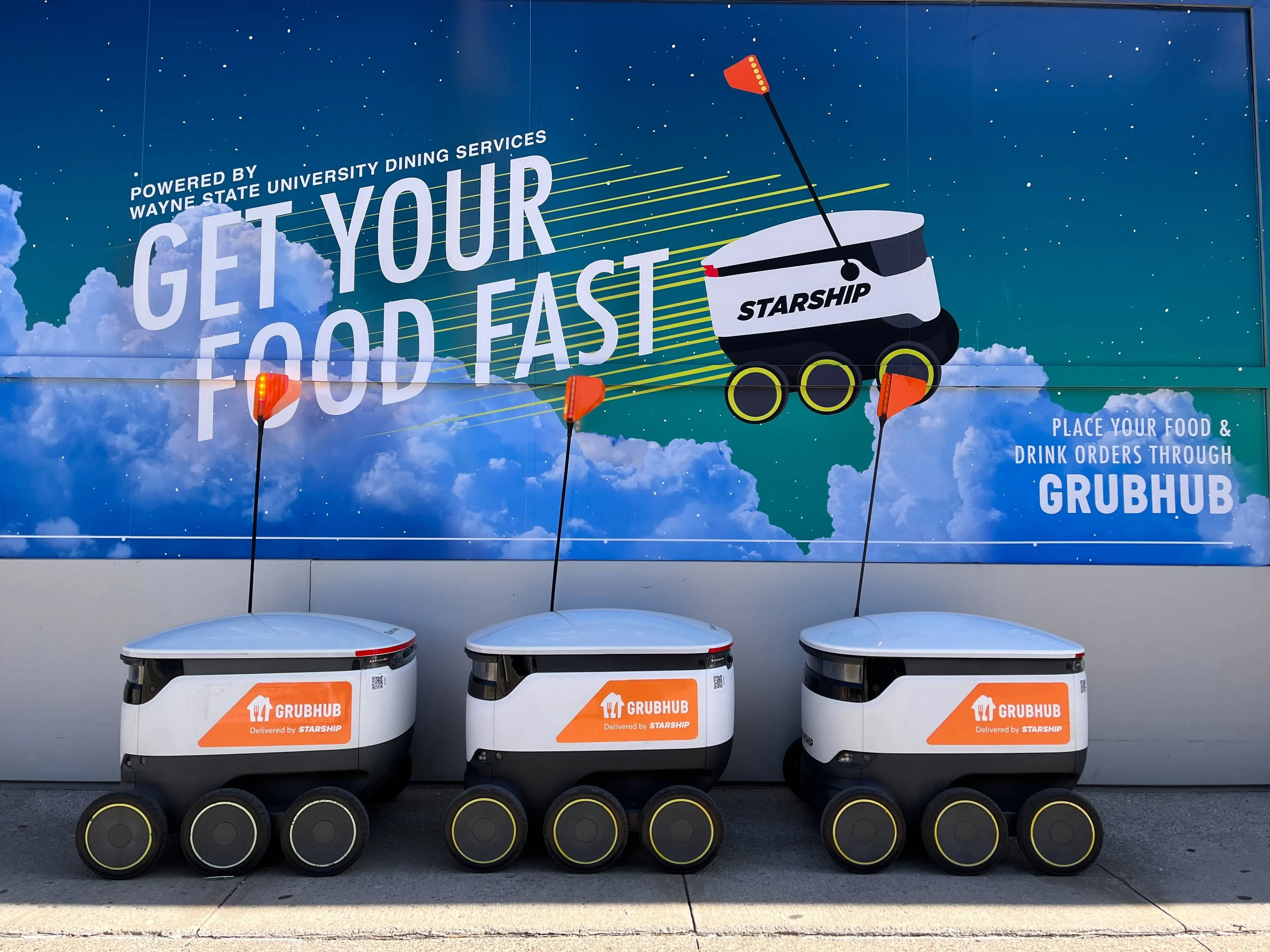 Three Starship sidewalk delivery robots with Grubhub logo under sign that says 'get your food fast'