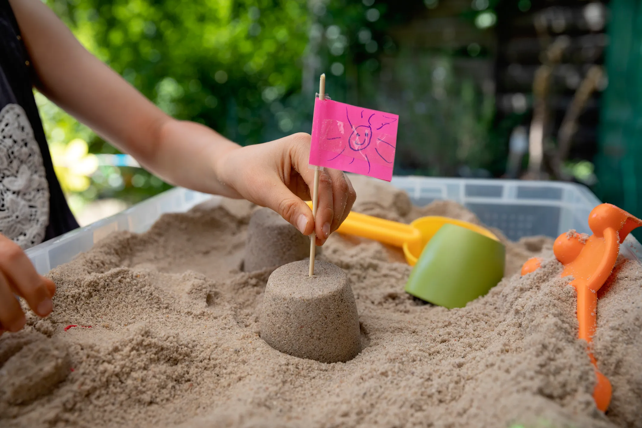 Child placing a homemade flag made from a pink sticky note with a drawing of a smiley face, into the top of a small sandcastle surrounded by sand inside a plastic sandbox.
