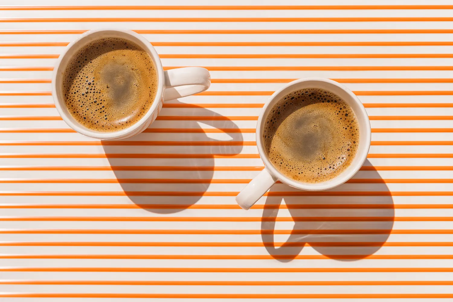 Two white coffee cup on orange striped table at direct sunlight. Top view with shadow.