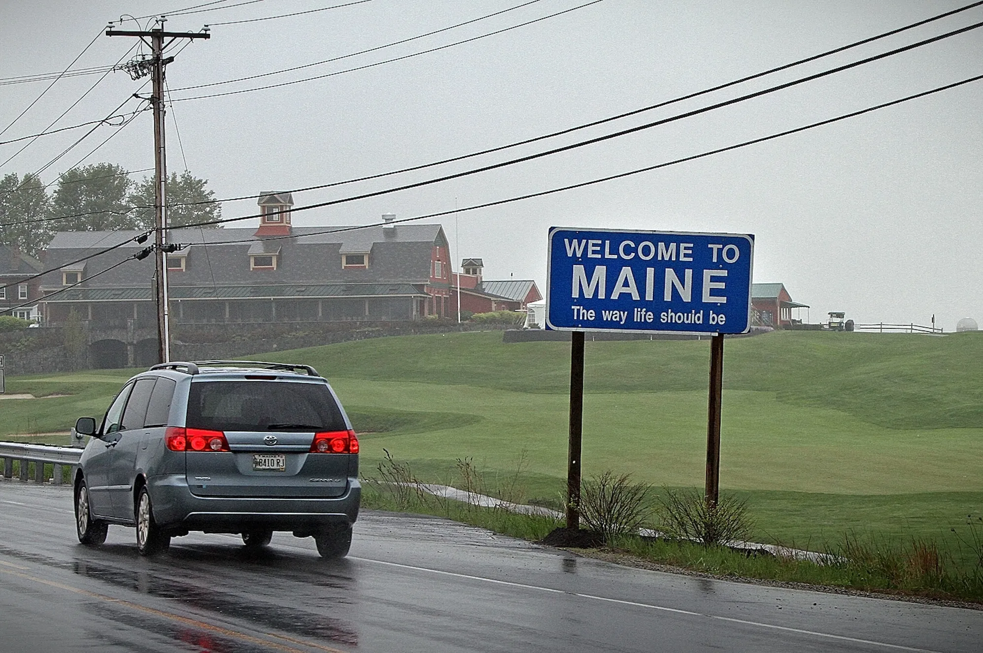 a blue roadsign that says "Welcome to Maine, the way life should be" on a road with a car driving past, on a dark and slightly rainy day with a large field and house in the background.