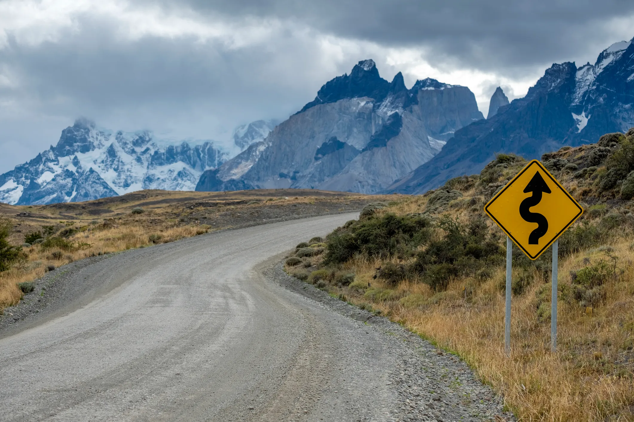 A gravel road with a warning of curves ahead as you head into a mountainous area with clouds above the mountains.