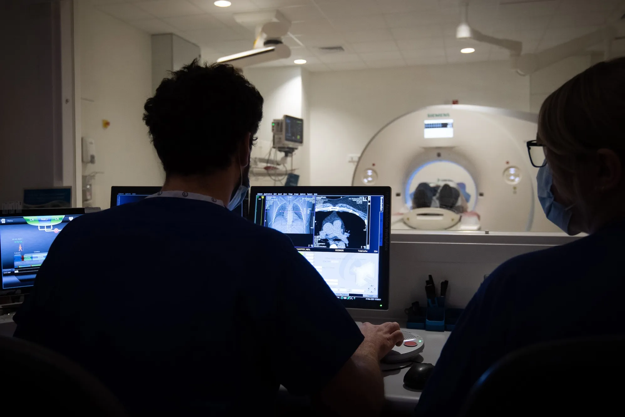 a photo of a person having a CT-scan in the background, with two radiographers behind protective glass in front of the CT scanner's computer
