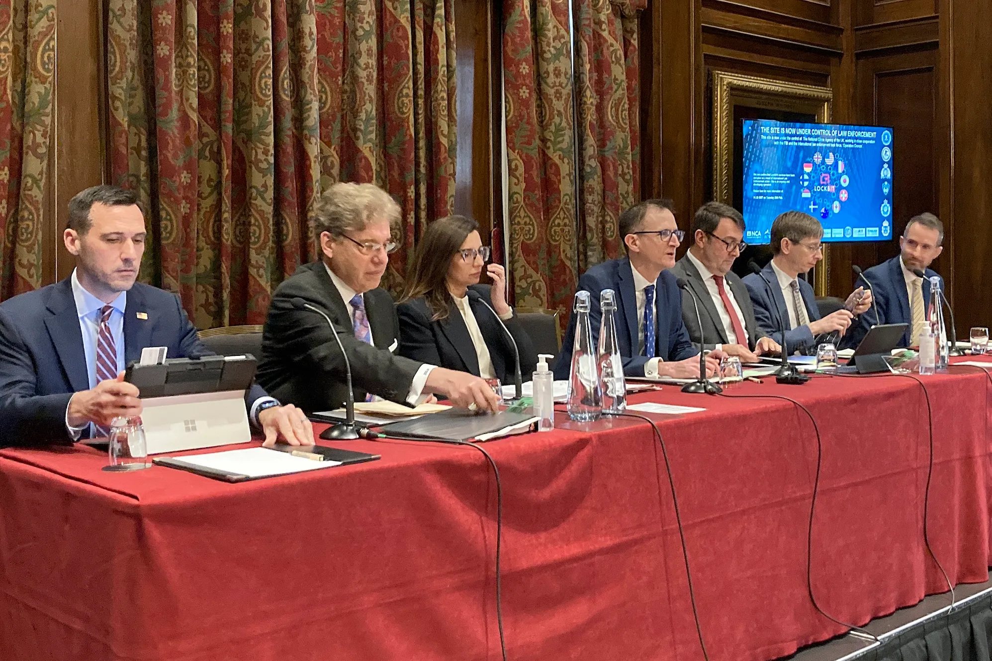 a photo of UK and US officials sitting at a red table discussing the LockBit takedown
