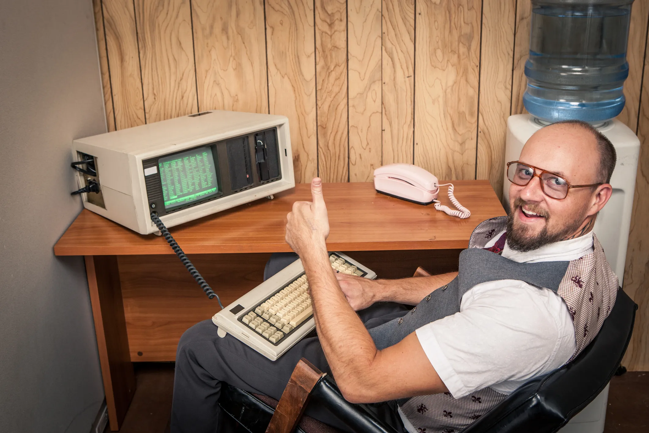 Happy office worker sitting, typing, working, and selling big at 1970's or 1980's monochrome computer. Retro office interior scene. Wood Paneling and a water cooler in the background. Old retro vintage technology