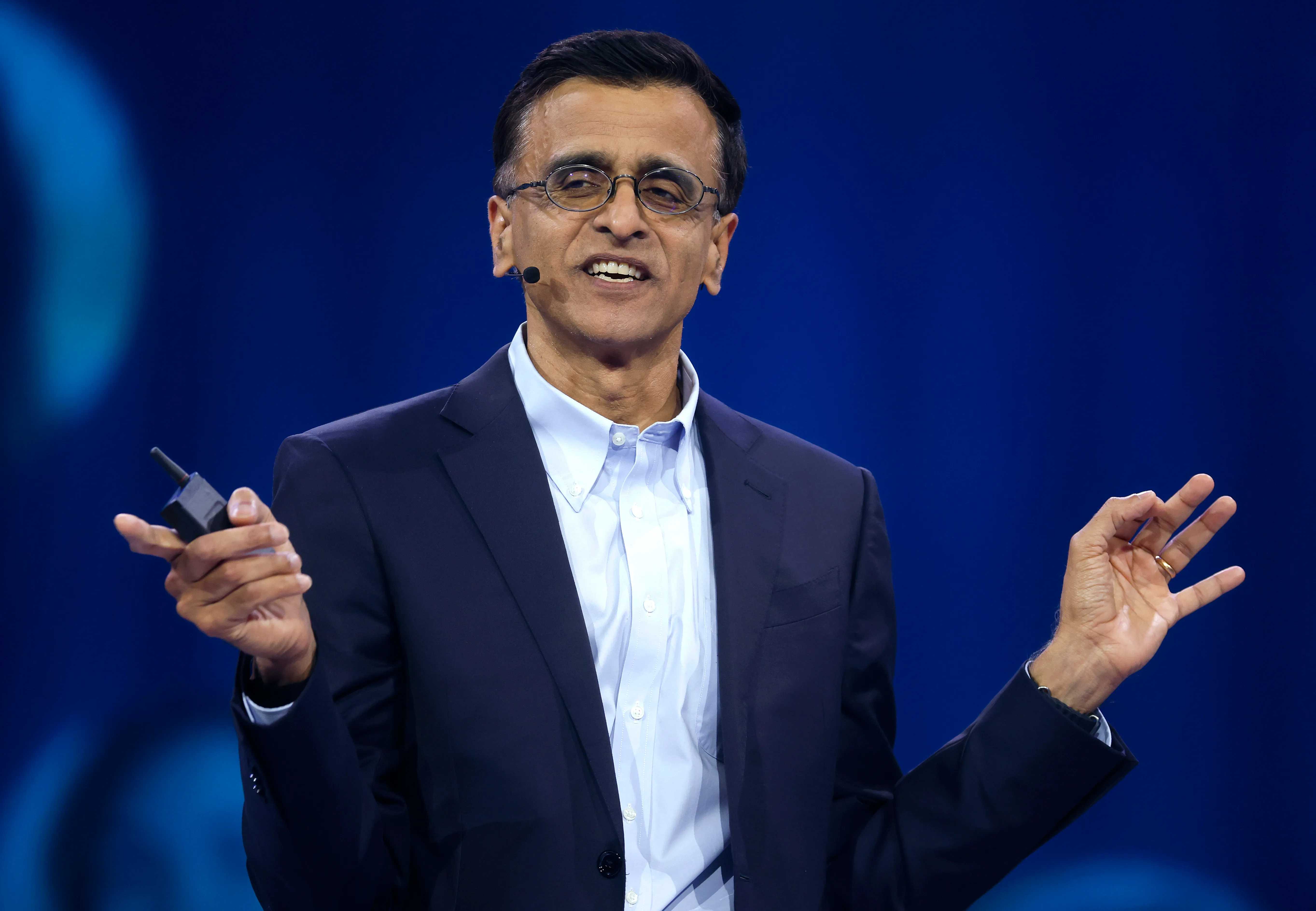 Snowflake CEO Sridhar Ramaswamy in a dark suit and light blue shirt speaks on stage against a blue background, wearing glasses and a headset microphone while gesturing with both hands.
